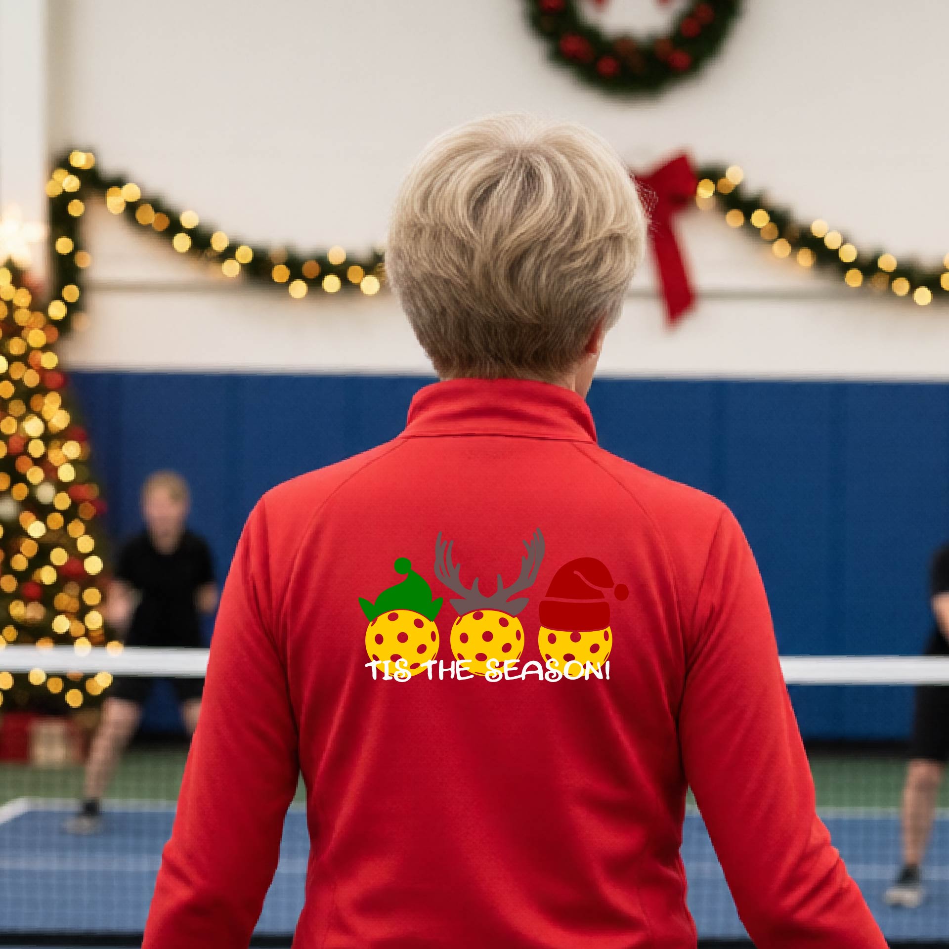 A woman viewed from behind wearing a red long-sleeve athletic top with a Christmas-themed graphic on the back. The graphic shows three yellow picKlebalLS with green, brown, and red holiday patterns, along with white text reading "TIS THE SEASON"
