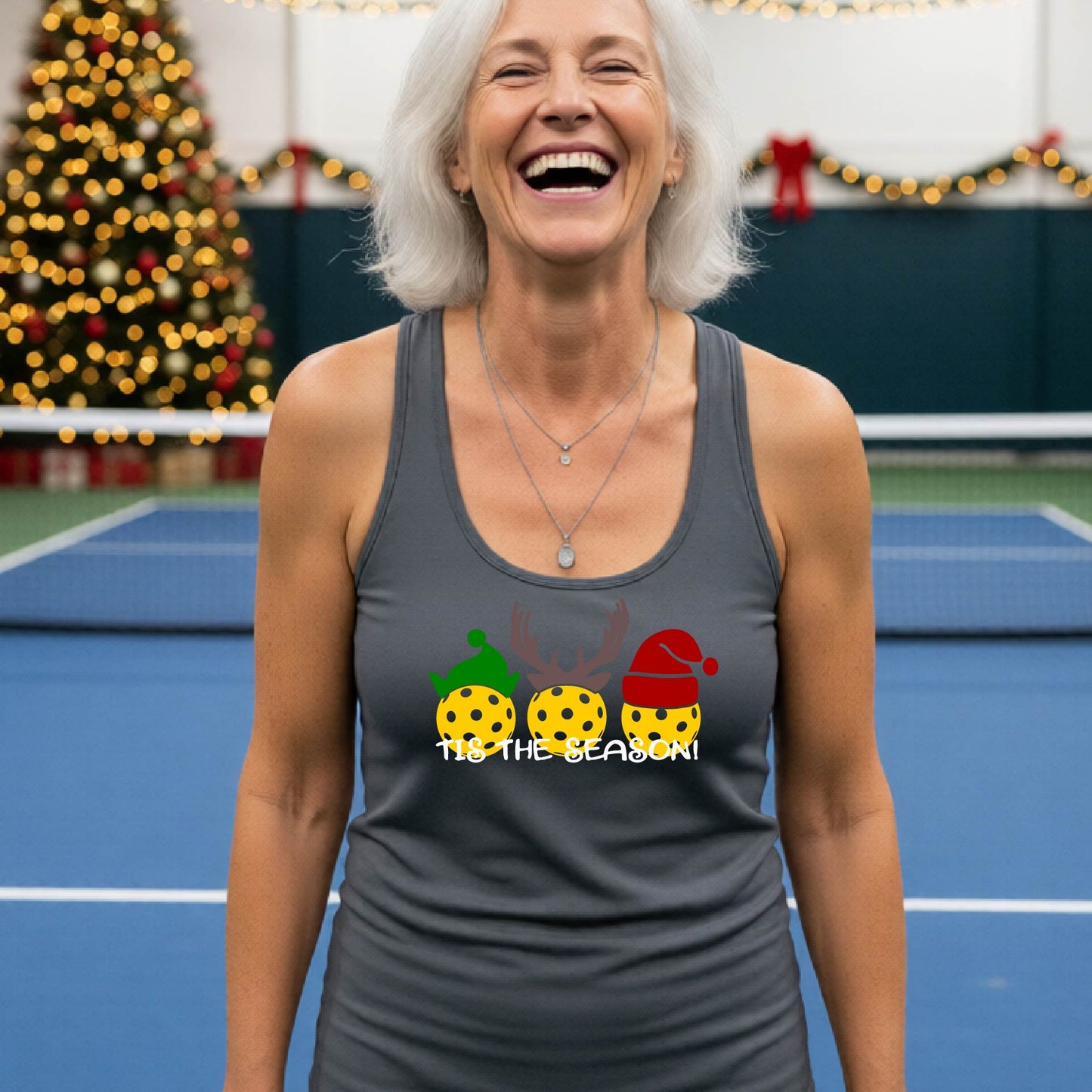 A woman laughing on an indoor pickleball court wearing a dark gray racerback tank top with a Christmas-themed graphic of three decorated pickleballs and the text 'TIS THE SEASON!'.