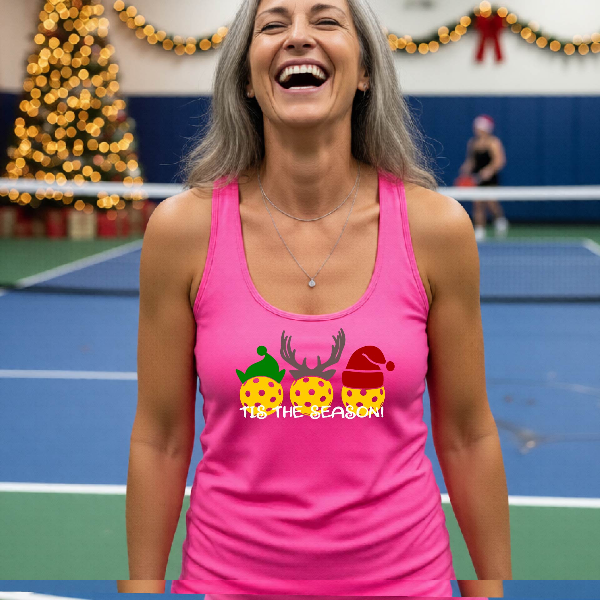 A woman laughing on an indoor pickleball court wearing a bright pink racerback tank top with a Christmas-themed graphic of three decorated pickleballs and the text 'TIS THE SEASON!'.