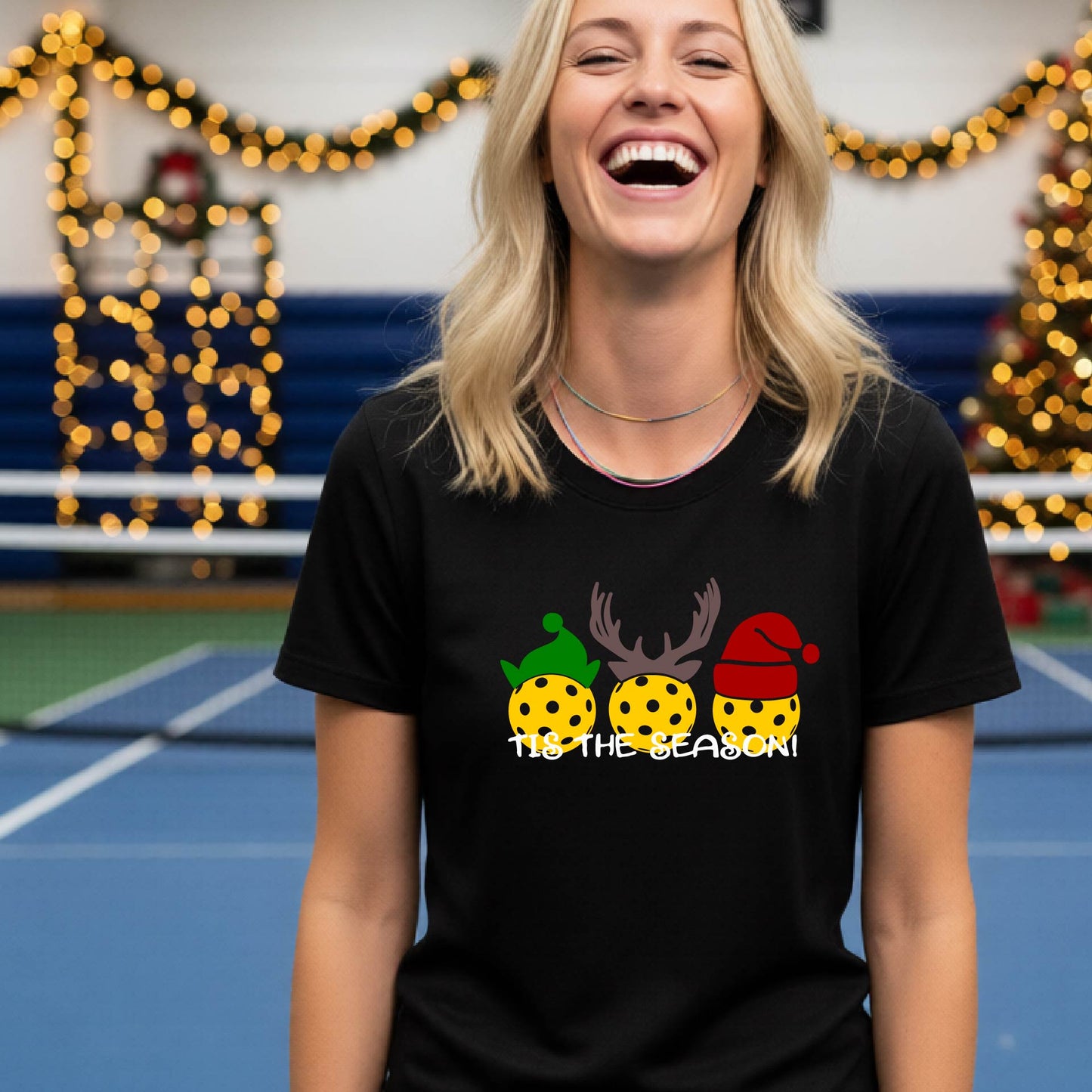 A woman laughing while wearing a black t-shirt with a Christmas graphic that reads 'TIS THE SEASON!' featuring three decorated pickleball and Christmas hats.. The background shows a holiday-themed indoor setting with Christmas tree lights, and stockings.