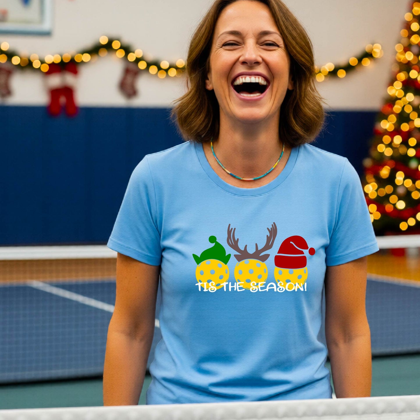 A woman laughing while wearing a light blue t-shirt with a Christmas graphic that reads 'TIS THE SEASON!' featuring three decorated pickleball and Christmas hats.. The background shows a holiday-themed indoor setting with Christmas tree lights, and stockings.