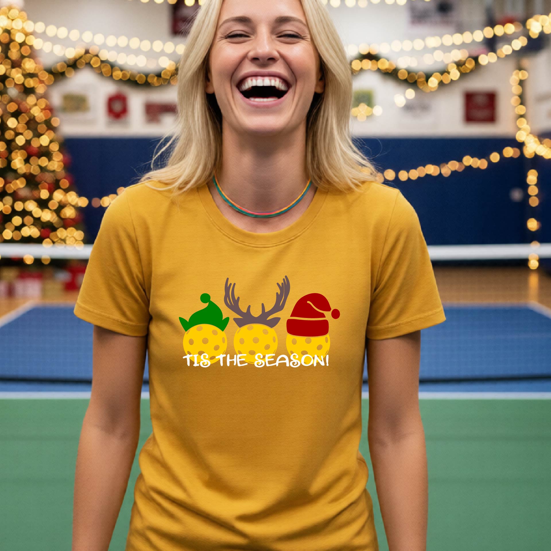 A woman laughing while wearing a mustard yellow gold t-shirt with a Christmas graphic that reads 'TIS THE SEASON!' featuring three decorated pickleball and Christmas hats.. The background shows a holiday-themed indoor setting with Christmas tree lights, and stockings.