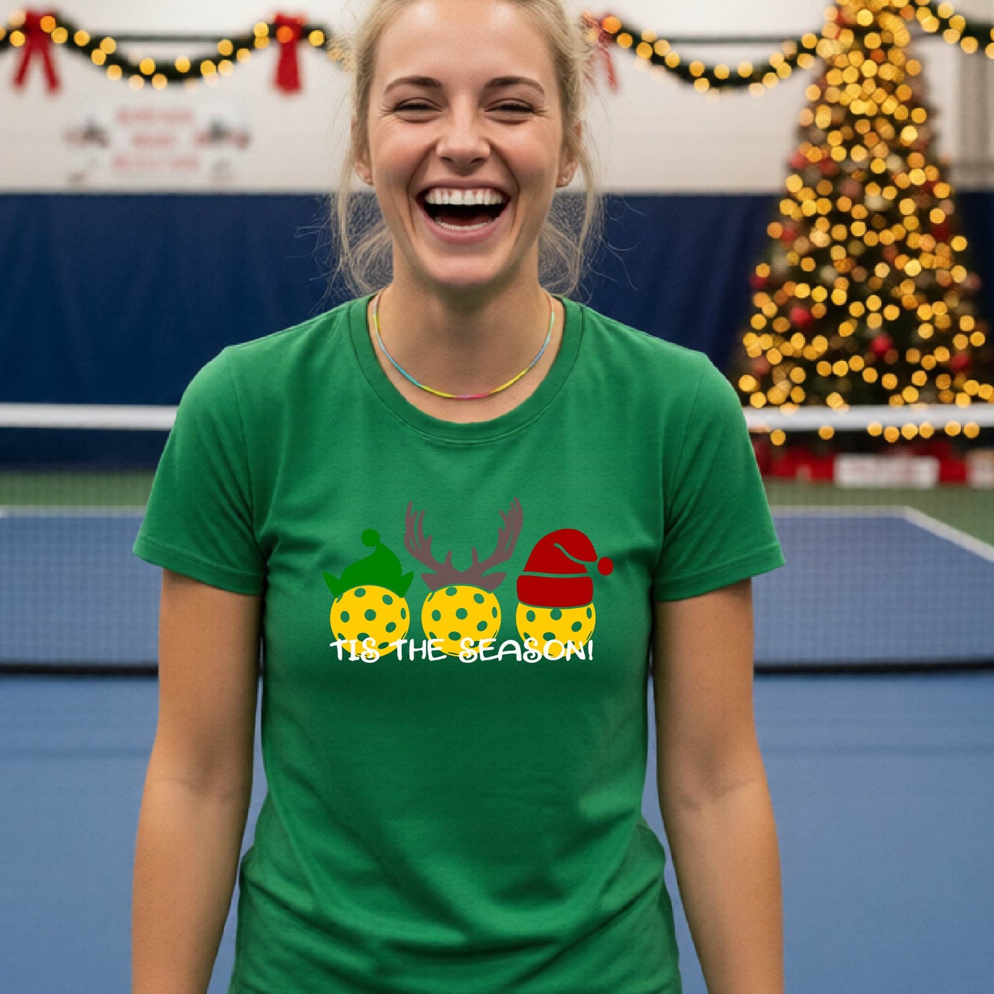 A woman laughing while wearing a green t-shirt with a Christmas graphic that reads 'TIS THE SEASON!' featuring three decorated pickleball and Christmas hats.. The background shows a holiday-themed indoor setting with Christmas tree lights, and stockings.