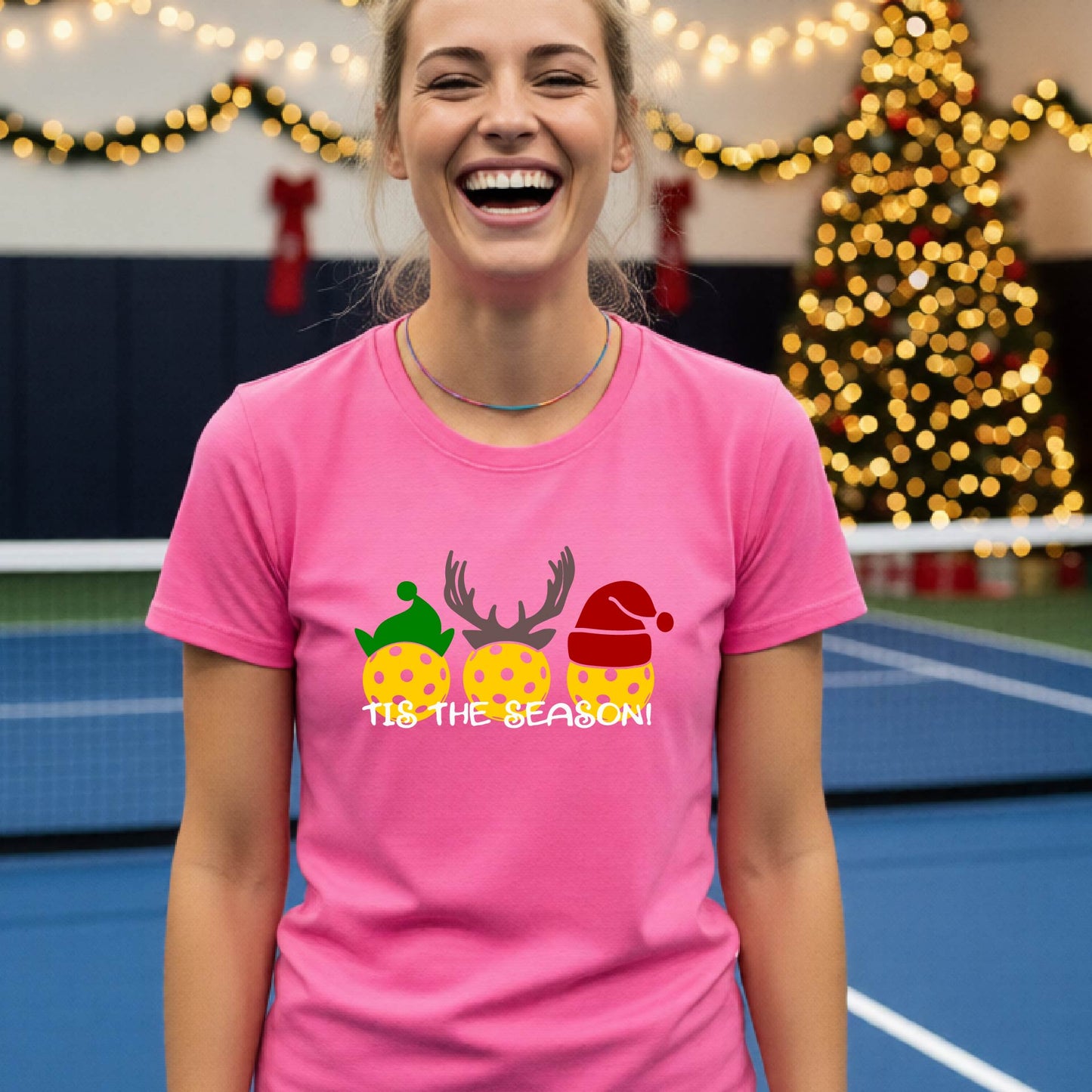 A woman laughing while wearing a bright piink shirt with a Christmas graphic that reads 'TIS THE SEASON!' featuring three decorated pickleball and Christmas hats.. The background shows a holiday-themed indoor setting with Christmas tree lights, and stockings.