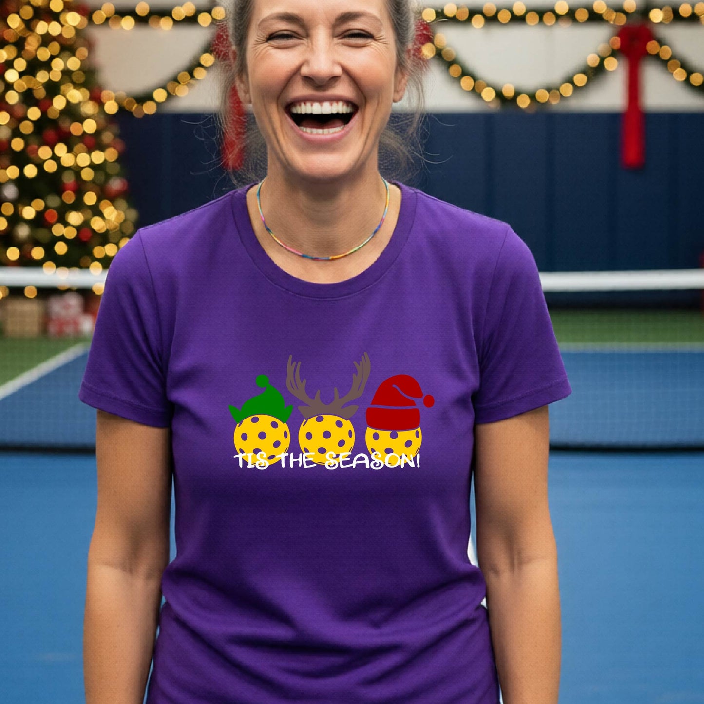 A woman laughing while wearing a purple t-shirt with a Christmas graphic that reads 'TIS THE SEASON!' featuring three decorated pickleball and Christmas hats.. The background shows a holiday-themed indoor setting with Christmas tree lights, and stockings.
