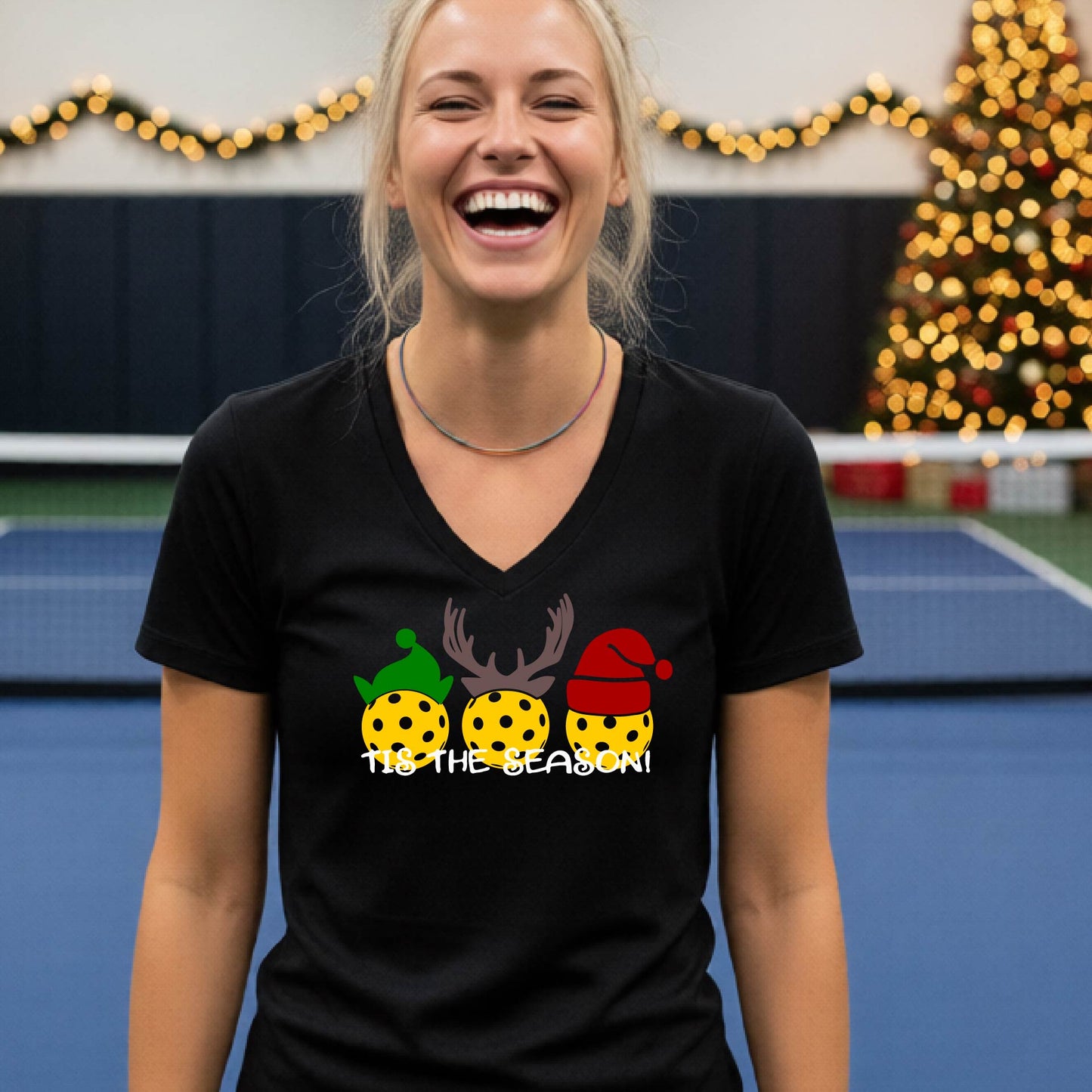 A woman laughing in a gym wearing a black v-neck t-shirt with a graphic of three holiday pickleballs wearing green, yellow, and red Santa hats, with the text 'TIE THE SEASON!' below them.