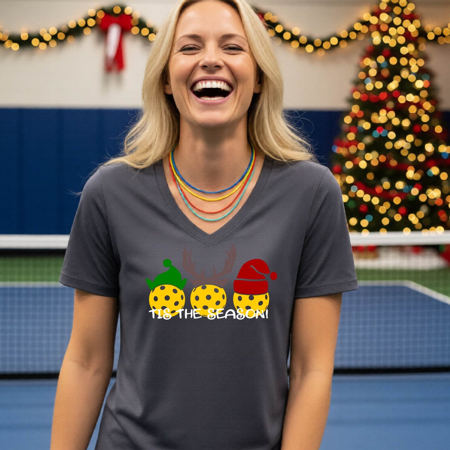 A woman laughing in a gym wearing a dark gray v-neck t-shirt with a graphic of three holiday pickleballs wearing green, yellow, and red Santa hats, with the text 'TIE THE SEASON!' below them.