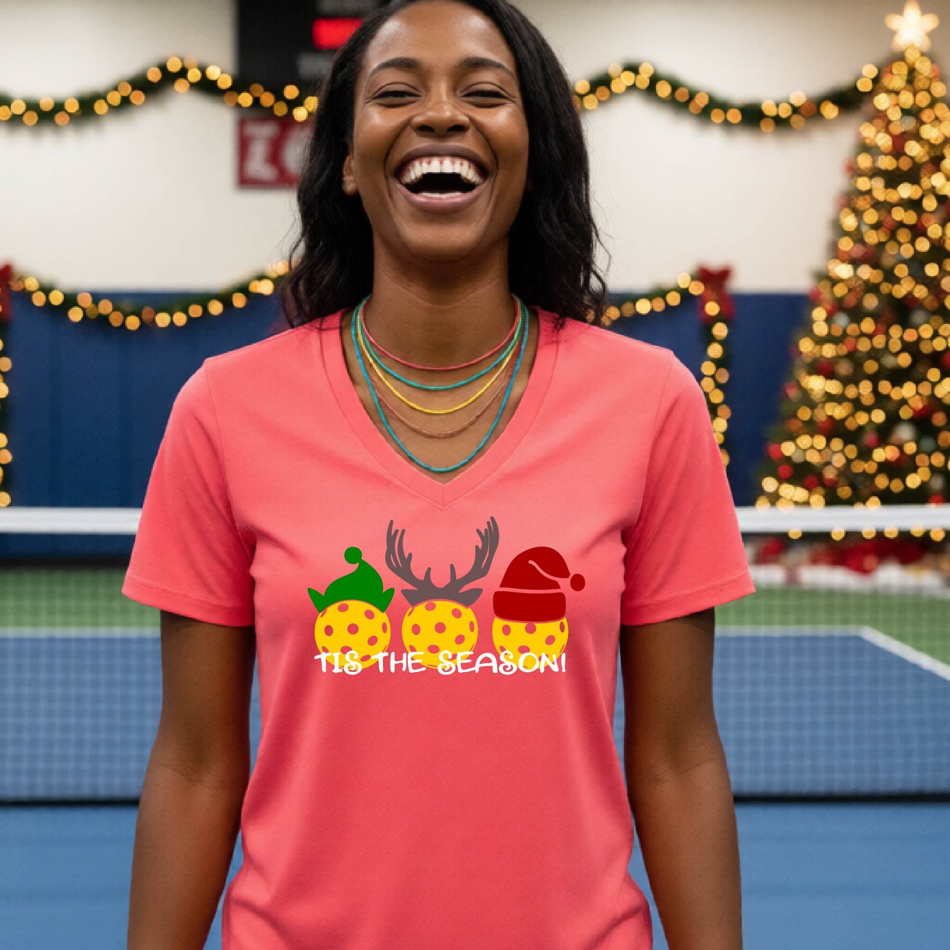 A woman laughing in a gym wearing a dark coral-pink v-neck t-shirt with a graphic of three holiday pickleballs wearing green, yellow, and red Santa hats, with the text 'TIE THE SEASON!' below them.