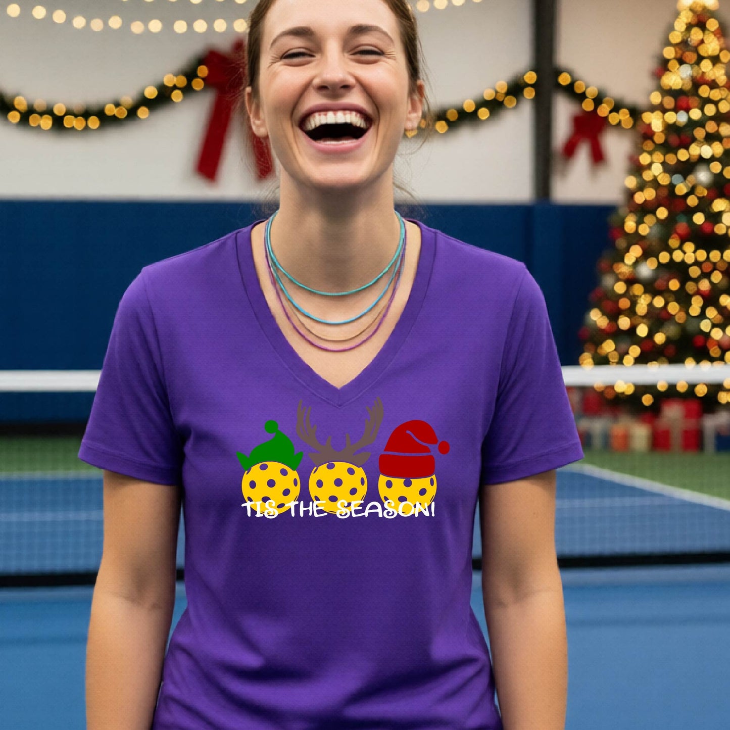 A woman laughing in a gym wearing a purple v-neck t-shirt with a graphic of three holiday pickleballs wearing green, yellow, and red Santa hats, with the text 'TIE THE SEASON!' below them.