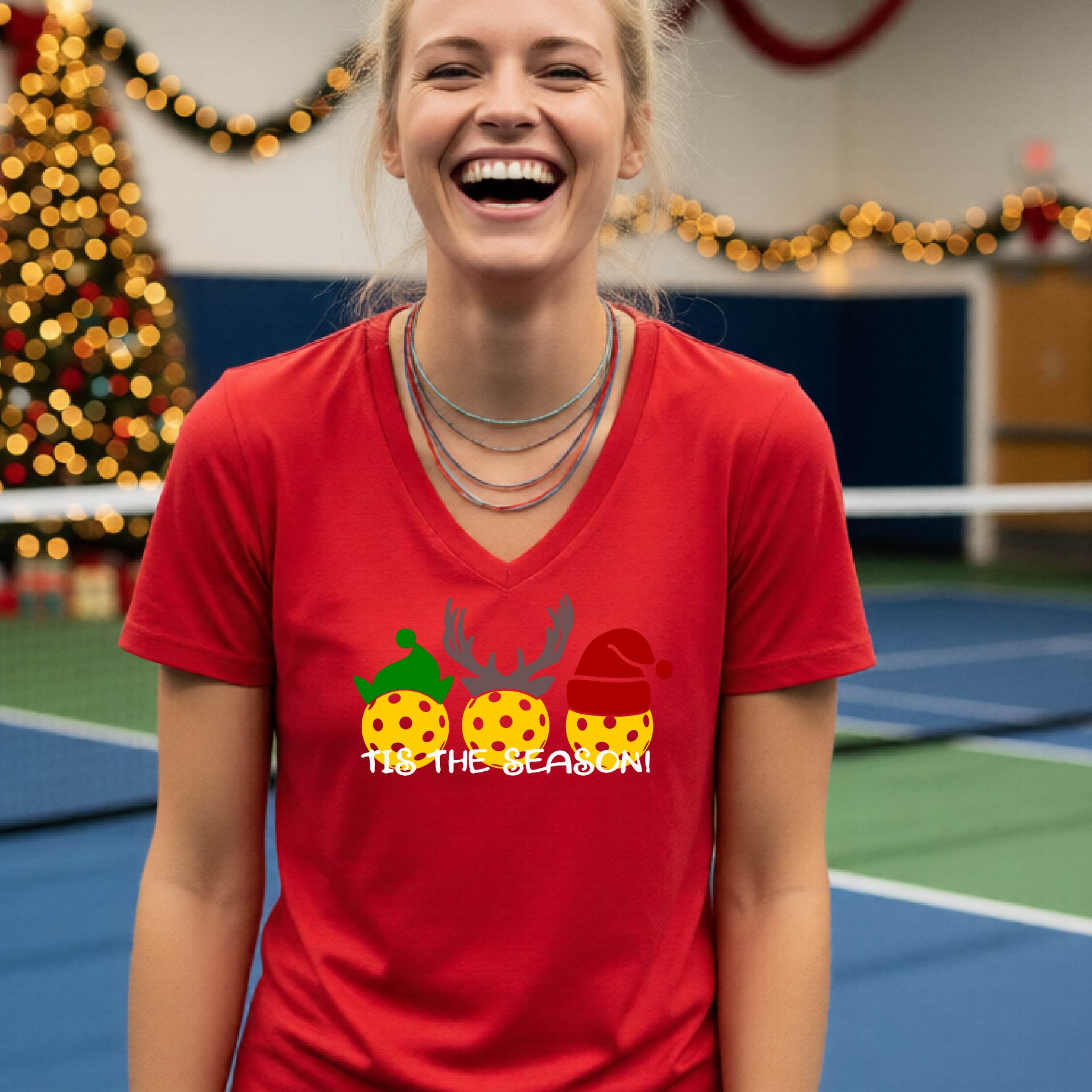 A woman laughing in a gym wearing a red v-neck t-shirt with a graphic of three holiday pickleballs wearing green, yellow, and red Santa hats, with the text 'TIE THE SEASON!' below them.