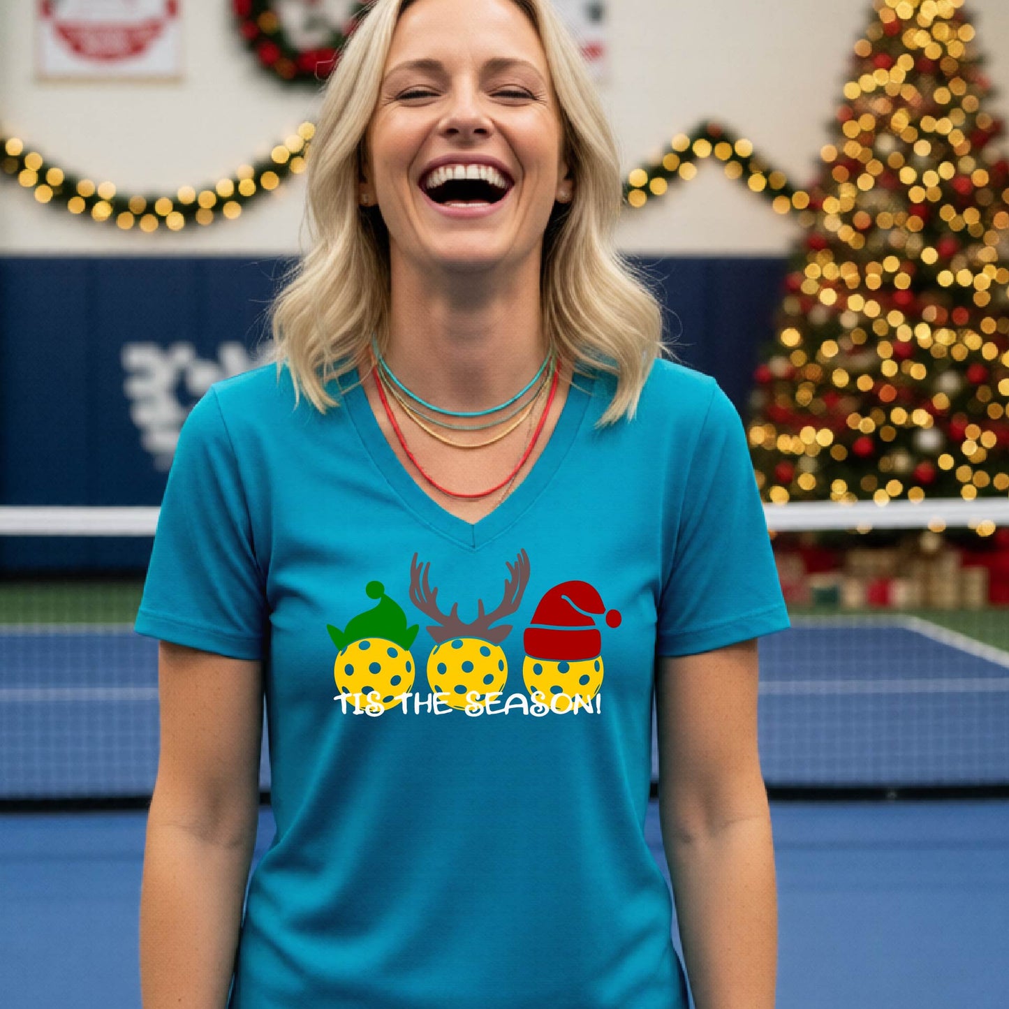A woman laughing in a gym wearing a teal v-neck t-shirt with a graphic of three holiday pickleballs wearing green, yellow, and red Santa hats, with the text 'TIE THE SEASON!' below them.