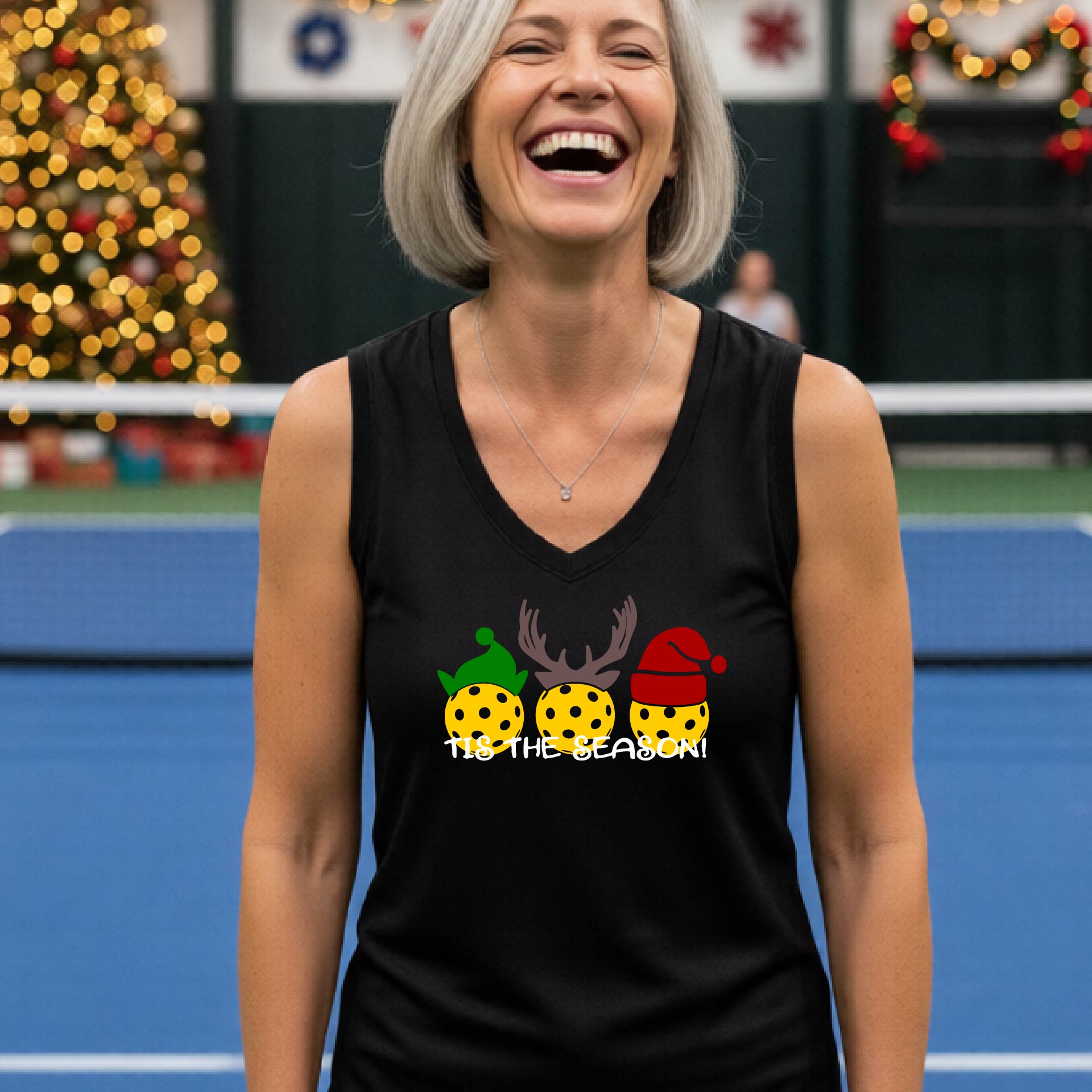 A woman with short gray hair smiling on a blue pickleball court, wearing a black sleeveless tank top with a holiday graphic of three yellow pickleballs wearing green, brown, and red hats, and the text 'TIS THE SEASON!'.
