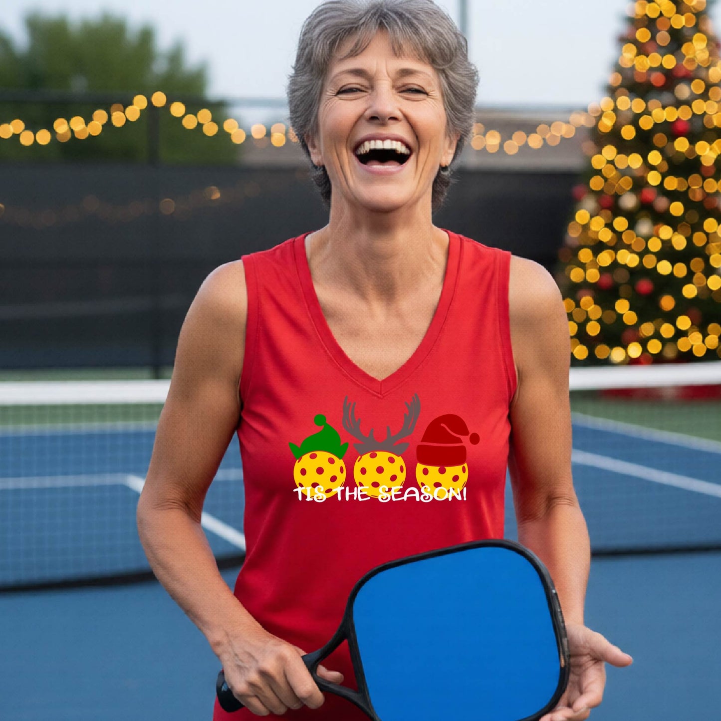A woman wsmiling on a blue pickleball court, wearing a red sleeveless tank top with a holiday graphic of three yellow pickleballs wearing green, brown, and red hats, and the text 'TIS THE SEASON!'.