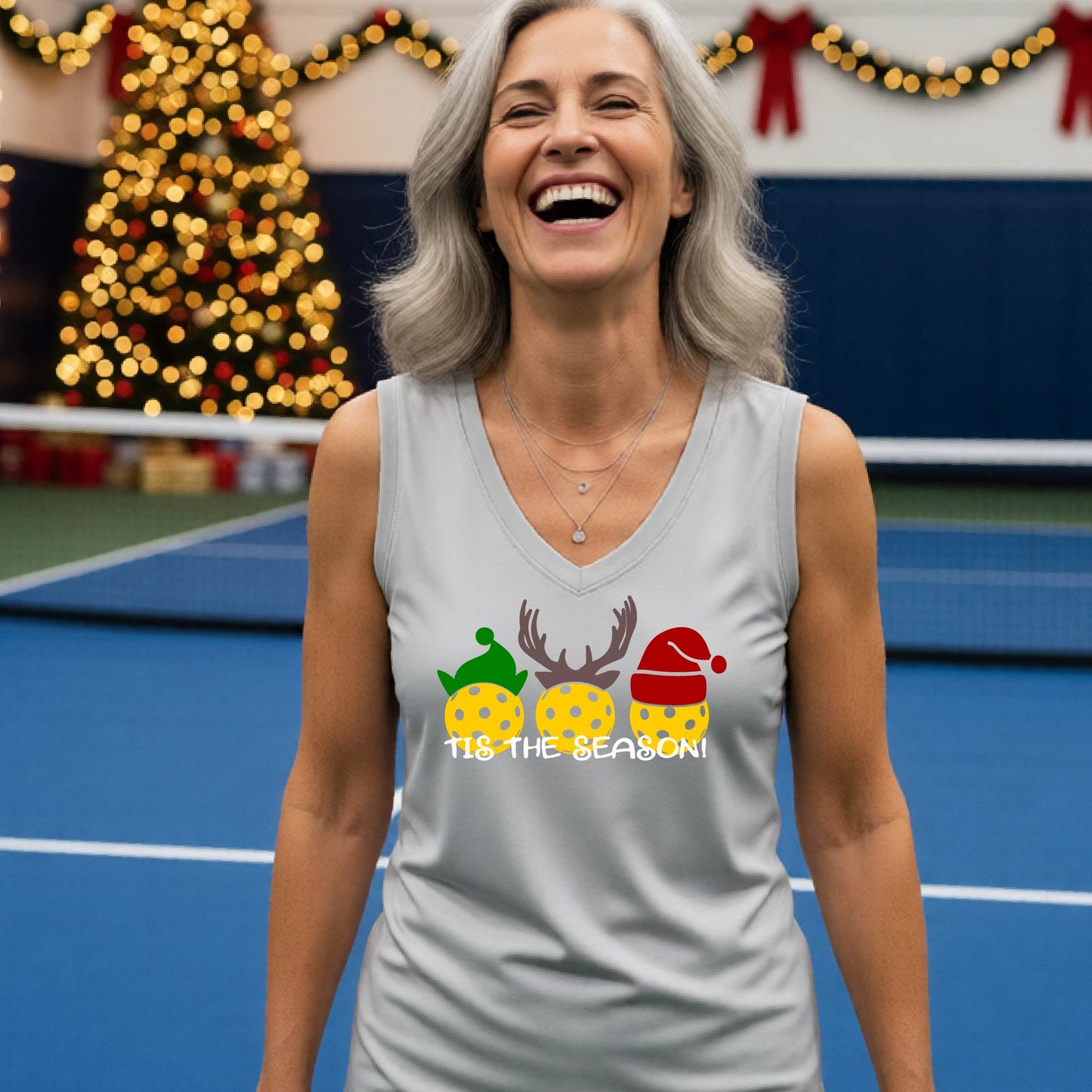 A woman smiling on a blue pickleball court, wearing a silver gray sleeveless tank top with a holiday graphic of three yellow pickleballs wearing green, brown, and red hats, and the text 'TIS THE SEASON!'.
