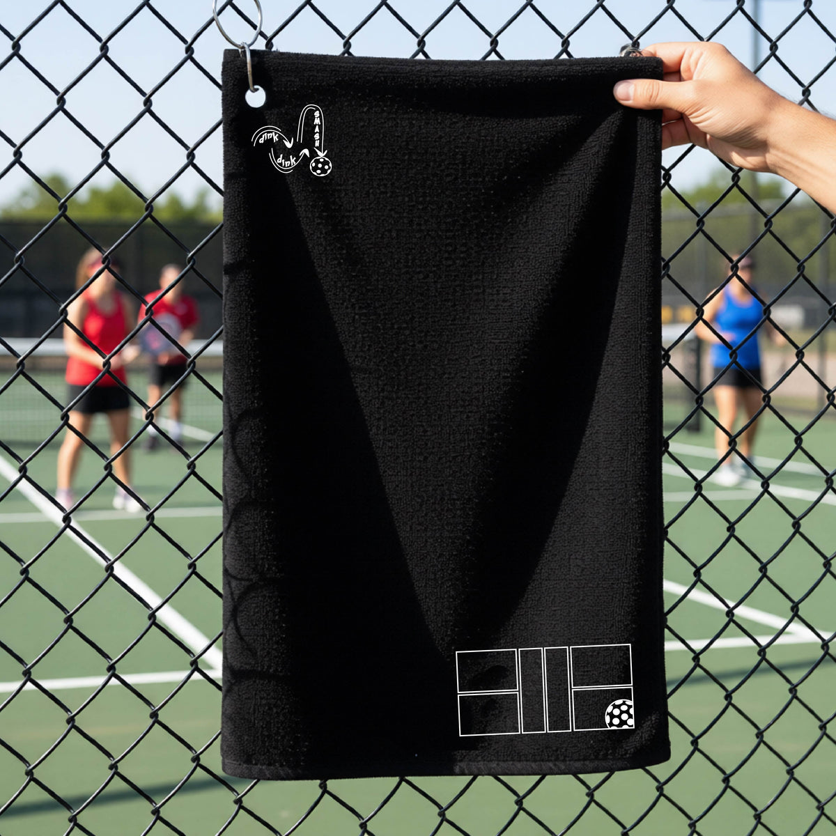 A close up of a black grommet and hook pickleball court towel with a DINK DINK SMASH graphic in the upper left corner and a white pickleball court with ball graphic in the bottom right corner. 