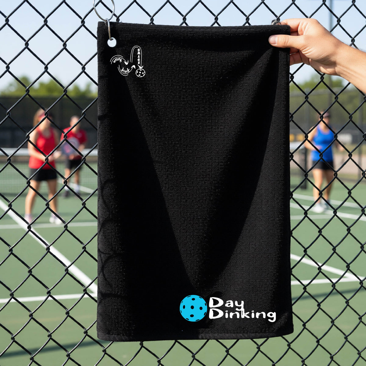 A black terry cloth towel held up by a hand in front of a chain-link fence and pickleball court, with the logo 'DINK DINK SMASH' in white on the top left and a colorful pickleball and the words "Day Dinking" printed near the bottom.
