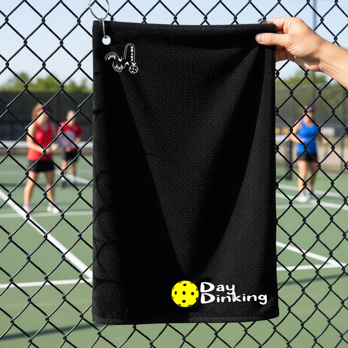 A black terry cloth towel held up by a hand in front of a chain-link fence and pickleball court, with the logo 'DINK DINK SMASH' in white on the top left and a colorful pickleball and the words "Day Dinking" printed near the bottom.