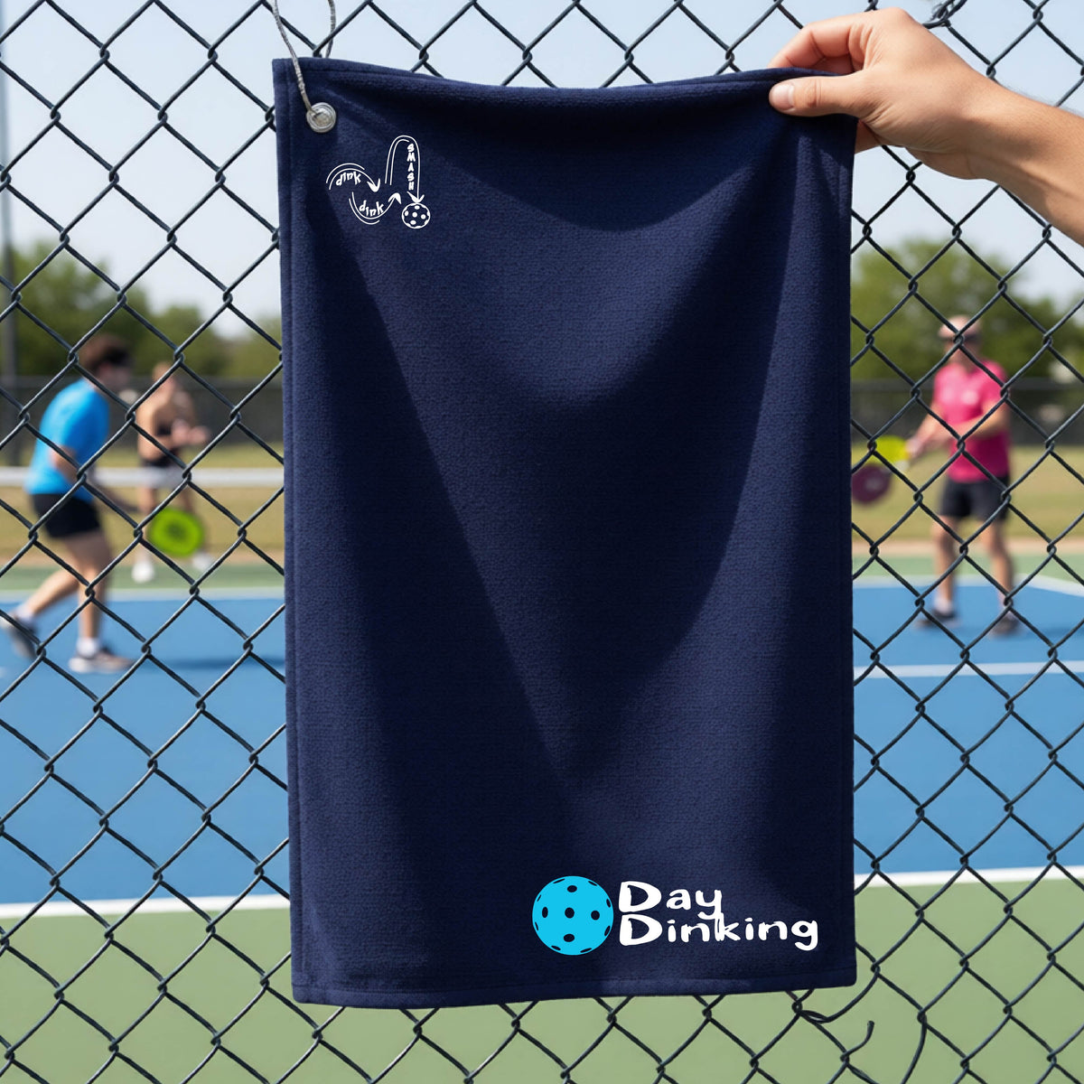 A navy blue terry cloth towel held up by a hand in front of a chain-link fence and pickleball court, with the logo 'DINK DINK SMASH' in white on the top left and a colorful pickleball and the words "Day Dinking" printed near the bottom.
