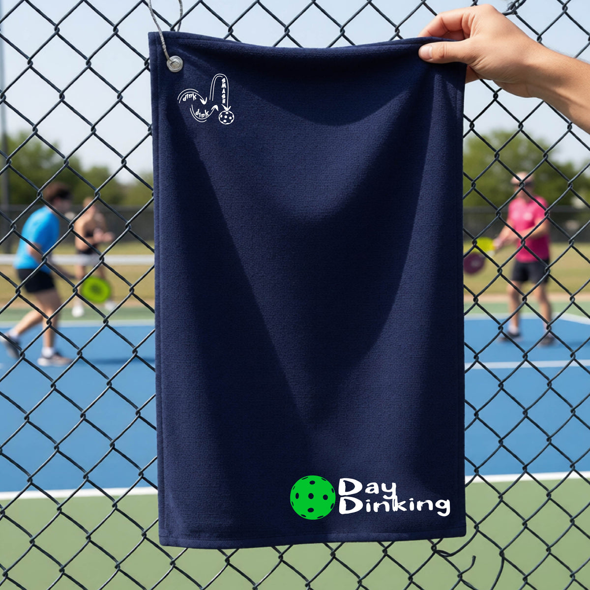 A navy blue terry cloth towel held up by a hand in front of a chain-link fence and pickleball court, with the logo 'DINK DINK SMASH' in white on the top left and a colorful pickleball and the words "Day Dinking" printed near the bottom.