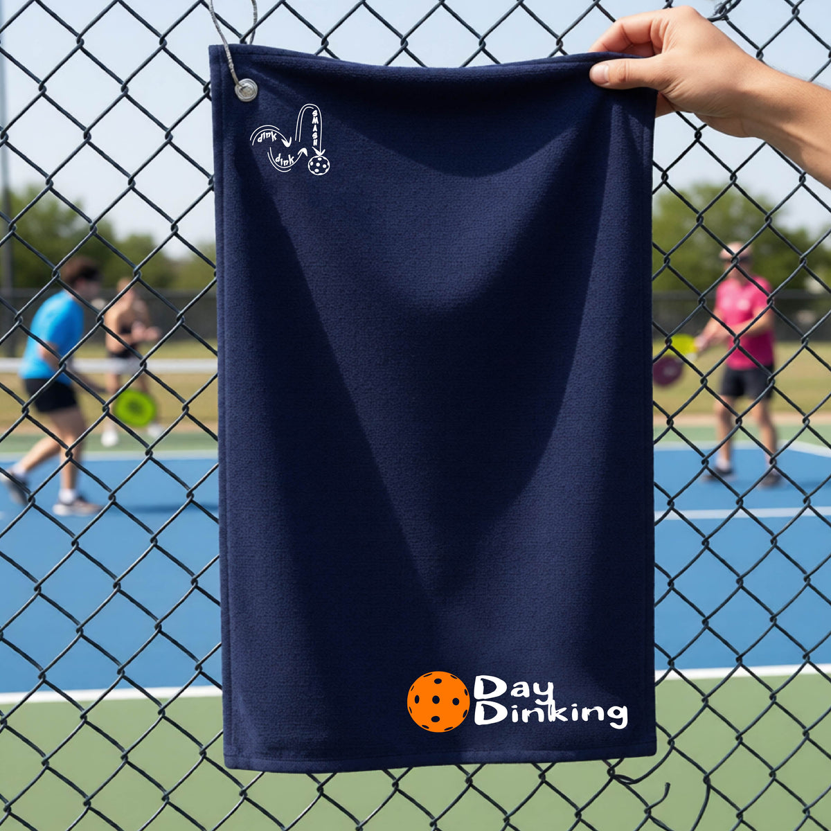 A navy blue terry cloth towel held up by a hand in front of a chain-link fence and pickleball court, with the logo 'DINK DINK SMASH' in white on the top left and a colorful pickleball and the words "Day Dinking" printed near the bottom.