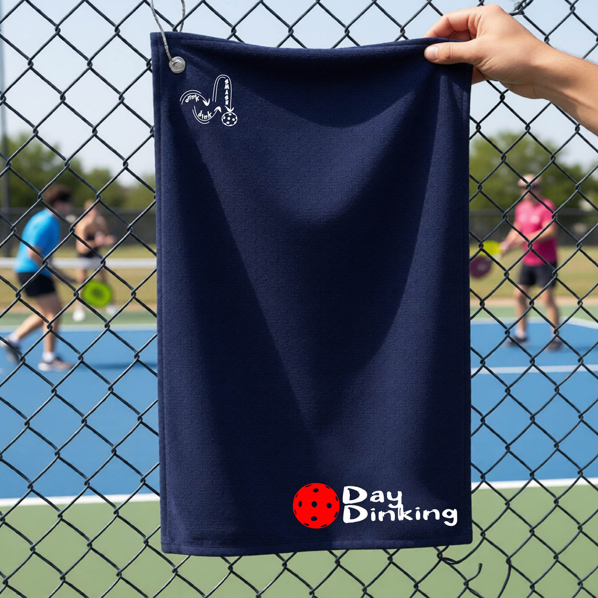 A navy blue terry cloth towel held up by a hand in front of a chain-link fence and pickleball court, with the logo 'DINK DINK SMASH' in white on the top left and a colorful pickleball and the words "Day Dinking" printed near the bottom.
