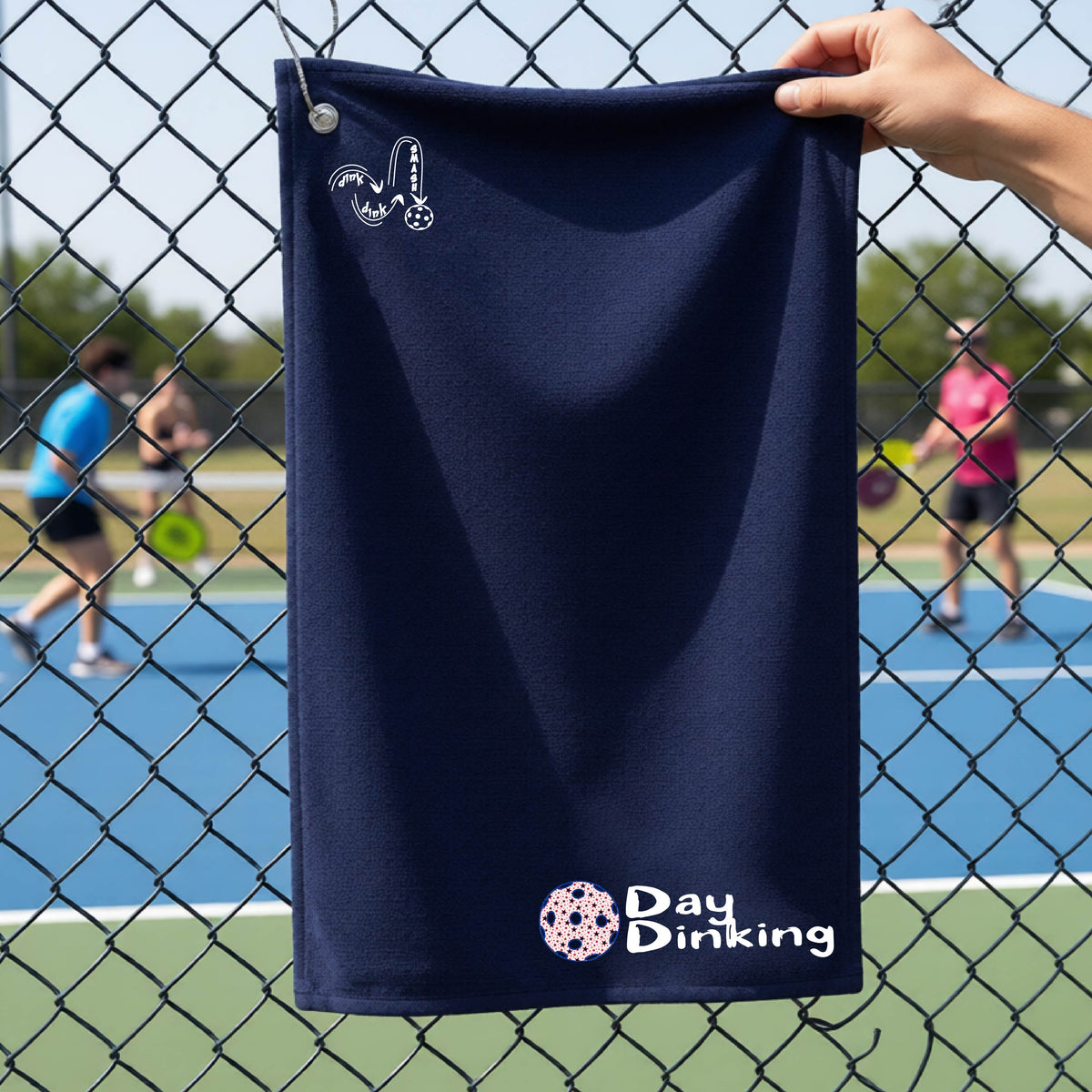 A navy blue terry cloth towel held up by a hand in front of a chain-link fence and pickleball court, with the logo 'DINK DINK SMASH' in white on the top left and a colorful pickleball and the words "Day Dinking" printed near the bottom.