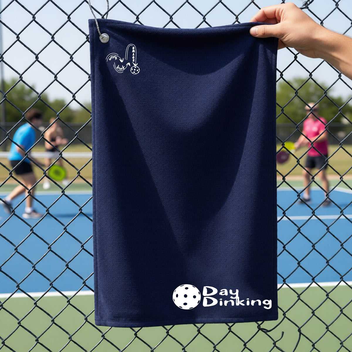 A navy blue terry cloth towel held up by a hand in front of a chain-link fence and pickleball court, with the logo 'DINK DINK SMASH' in white on the top left and a colorful pickleball and the words "Day Dinking" printed near the bottom.
