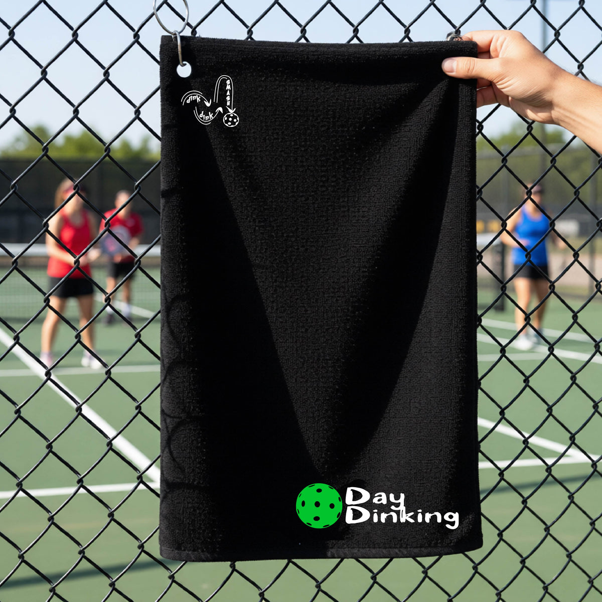 A black terry cloth towel held up by a hand in front of a chain-link fence and pickleball court, with the logo 'DINK DINK SMASH' in white on the top left and a colorful pickleball and the words "Day Dinking" printed near the bottom.