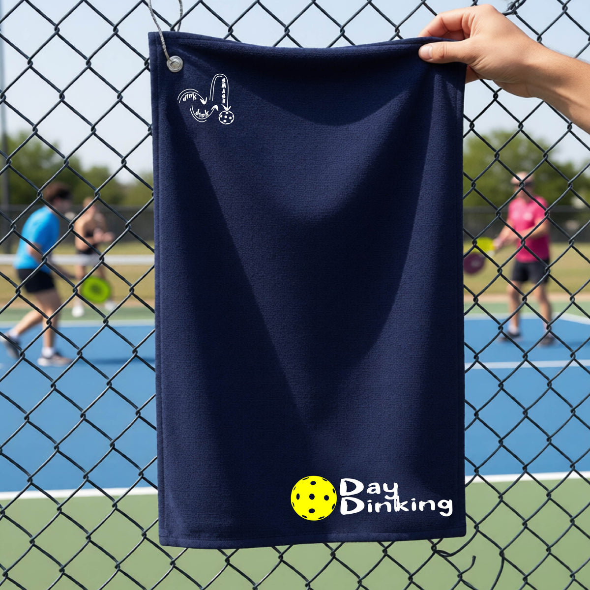 A navy blue terry cloth towel held up by a hand in front of a chain-link fence and pickleball court, with the logo 'DINK DINK SMASH' in white on the top left and a colorful pickleball and the words "Day Dinking" printed near the bottom.