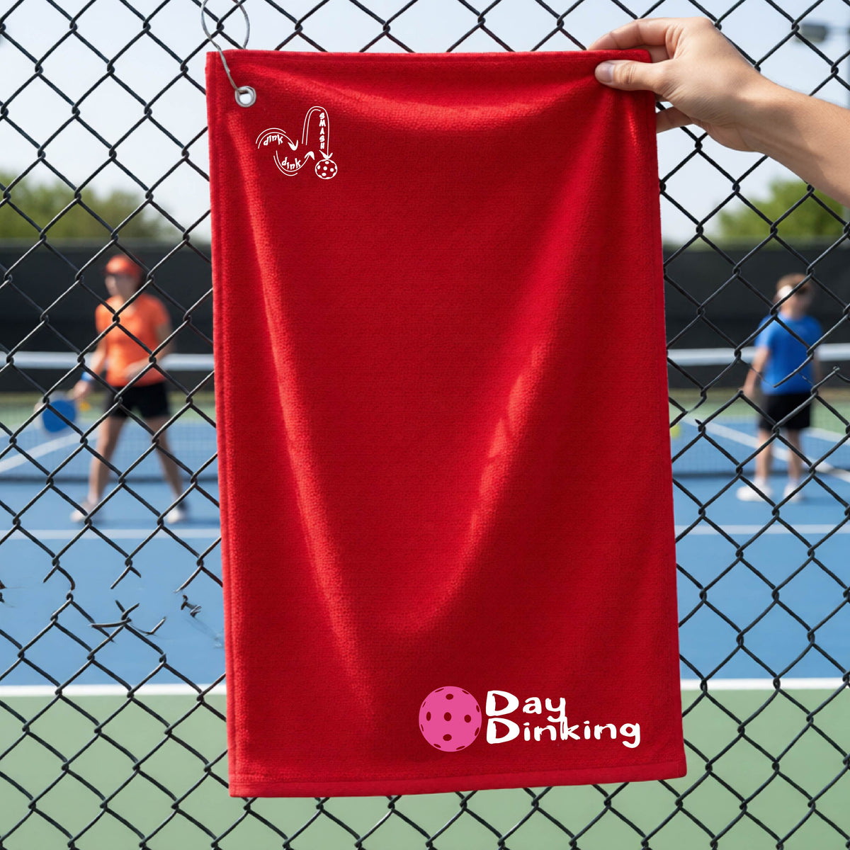A red terry cloth towel held up by a hand in front of a chain-link fence and pickleball court, with the logo 'DINK DINK SMASH' in white on the top left and a colorful pickleball and the words "Day Dinking" printed near the bottom.