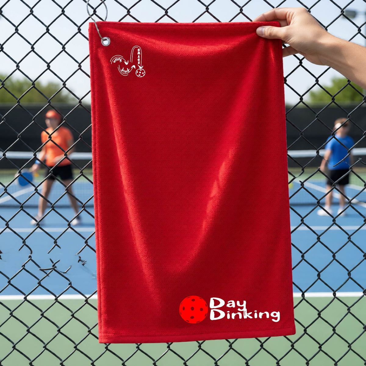 A red terry cloth towel held up by a hand in front of a chain-link fence and pickleball court, with the logo 'DINK DINK SMASH' in white on the top left and a colorful pickleball and the words "Day Dinking" printed near the bottom.