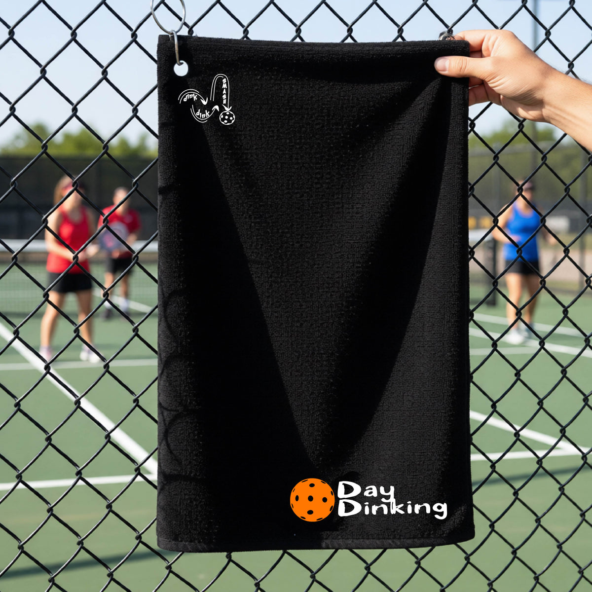A black terry cloth towel held up by a hand in front of a chain-link fence and pickleball court, with the logo 'DINK DINK SMASH' in white on the top left and a colorful pickleball and the words "Day Dinking" printed near the bottom.