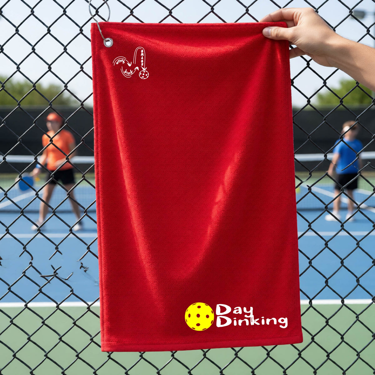A red terry cloth towel held up by a hand in front of a chain-link fence and pickleball court, with the logo 'DINK DINK SMASH' in white on the top left and a colorful pickleball and the words "Day Dinking" printed near the bottom.