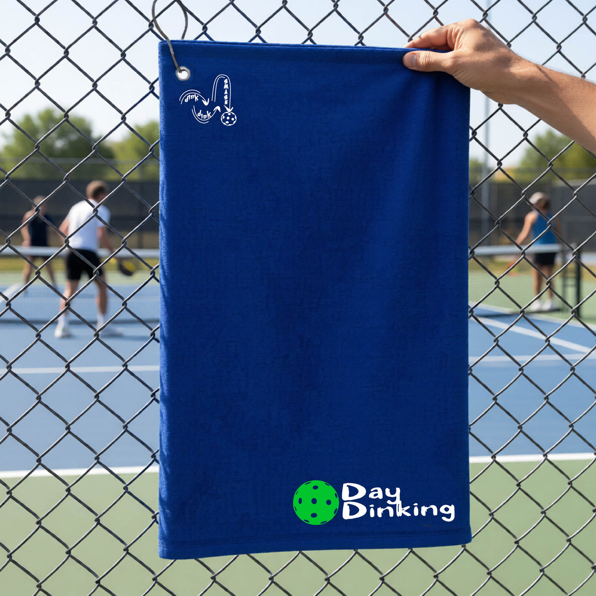 A royal blue terry cloth towel held up by a hand in front of a chain-link fence and pickleball court, with the logo 'DINK DINK SMASH' in white on the top left and a colorful pickleball and the words "Day Dinking" printed near the bottom.
