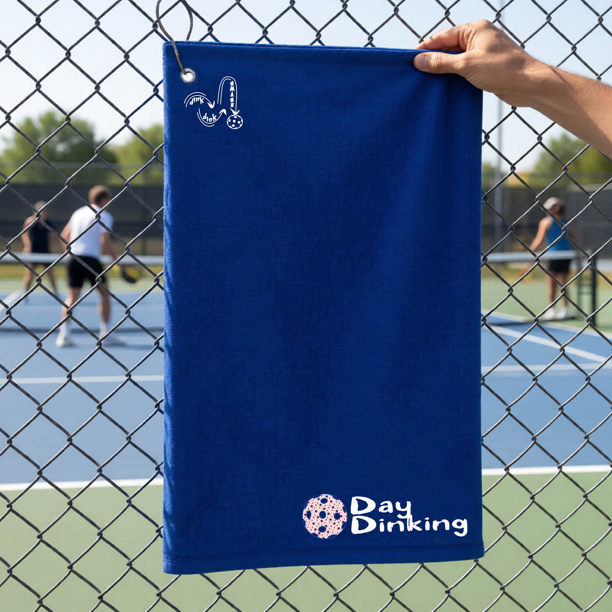 A royal blue terry cloth towel held up by a hand in front of a chain-link fence and pickleball court, with the logo 'DINK DINK SMASH' in white on the top left and a colorful pickleball and the words "Day Dinking" printed near the bottom.