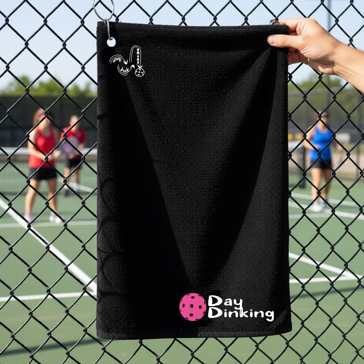 A black terry cloth towel held up by a hand in front of a chain-link fence and pickleball court, with the logo 'DINK DINK SMASH' in white on the top left and a colorful pickleball and the words "Day Dinking" printed near the bottom.