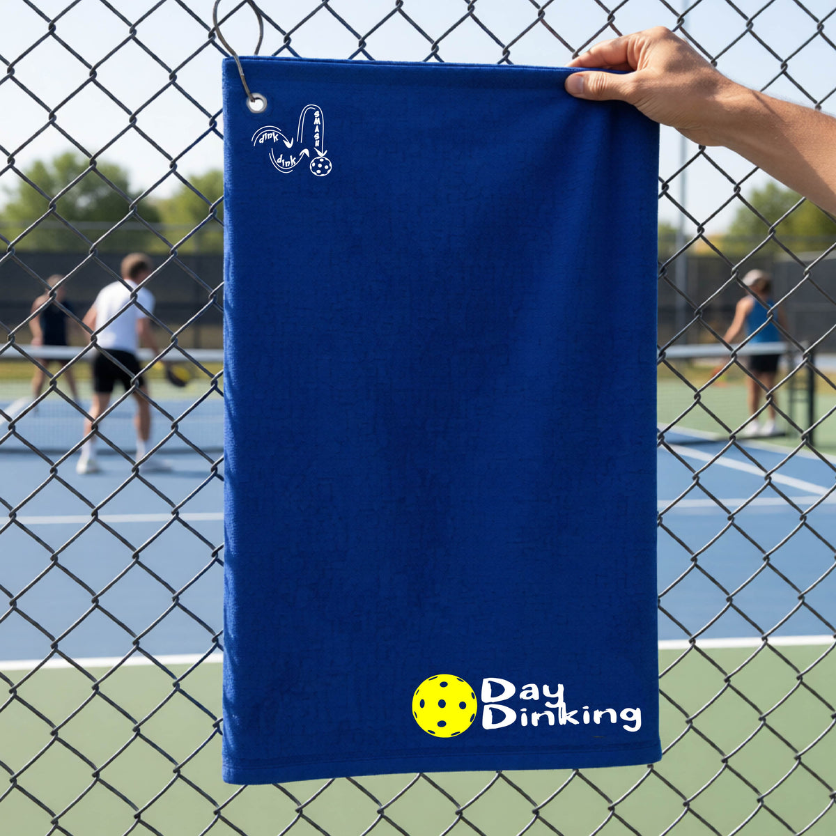 A royal blue terry cloth towel held up by a hand in front of a chain-link fence and pickleball court, with the logo 'DINK DINK SMASH' in white on the top left and a colorful pickleball and the words "Day Dinking" printed near the bottom.