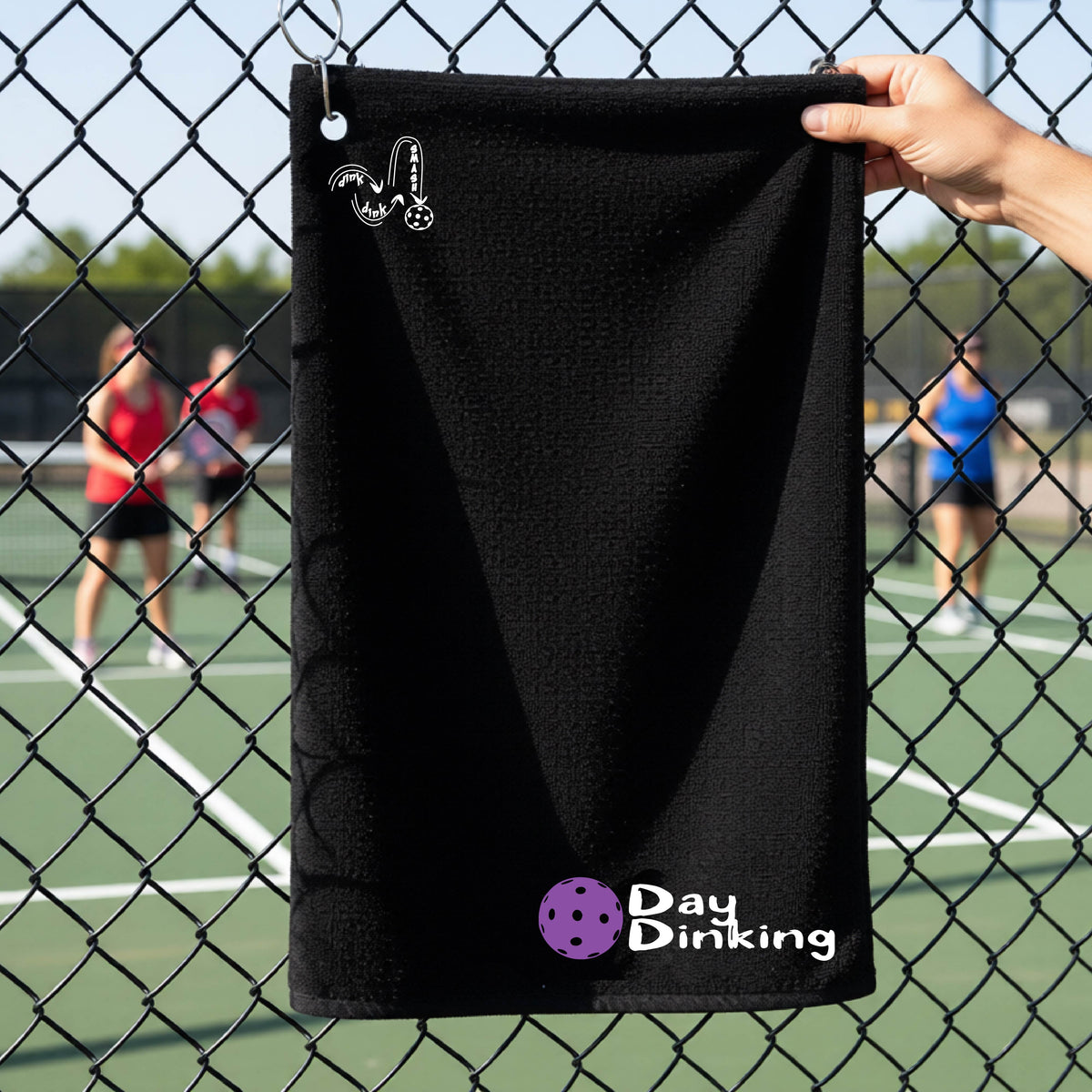 A black terry cloth towel held up by a hand in front of a chain-link fence and pickleball court, with the logo 'DINK DINK SMASH' in white on the top left and a colorful pickleball and the words "Day Dinking" printed near the bottom.