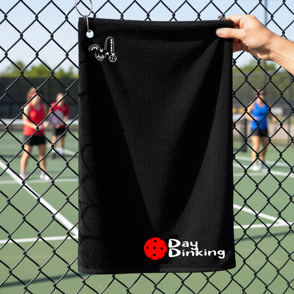 A black terry cloth towel held up by a hand in front of a chain-link fence and pickleball court, with the logo 'DINK DINK SMASH' in white on the top left and a colorful pickleball and the words "Day Dinking" printed near the bottom.