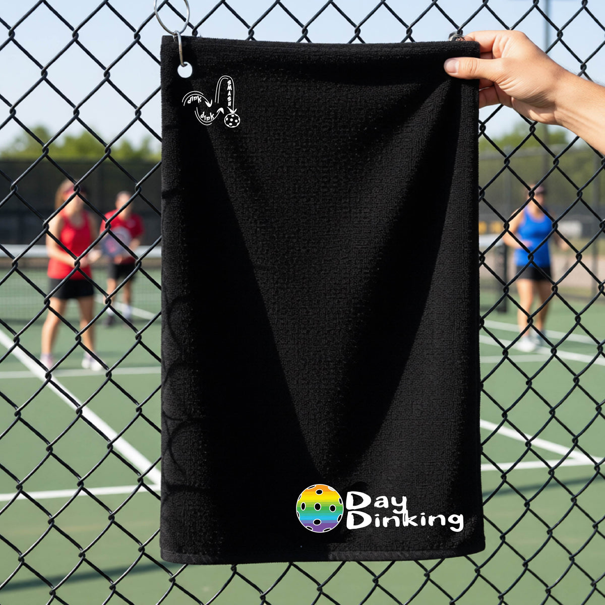 A black terry cloth towel held up by a hand in front of a chain-link fence and pickleball court, with the logo 'DINK DINK SMASH' in white on the top left and a colorful pickleball and the words "Day Dinking" printed near the bottom.
