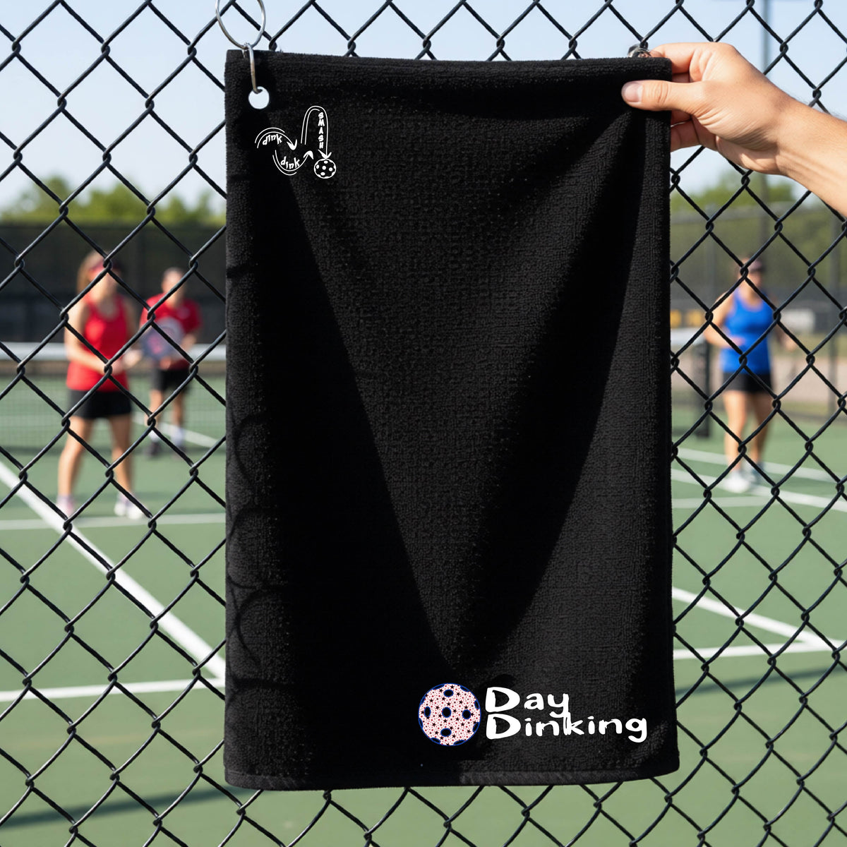 A black terry cloth towel held up by a hand in front of a chain-link fence and pickleball court, with the logo 'DINK DINK SMASH' in white on the top left and a colorful pickleball and the words "Day Dinking" printed near the bottom.