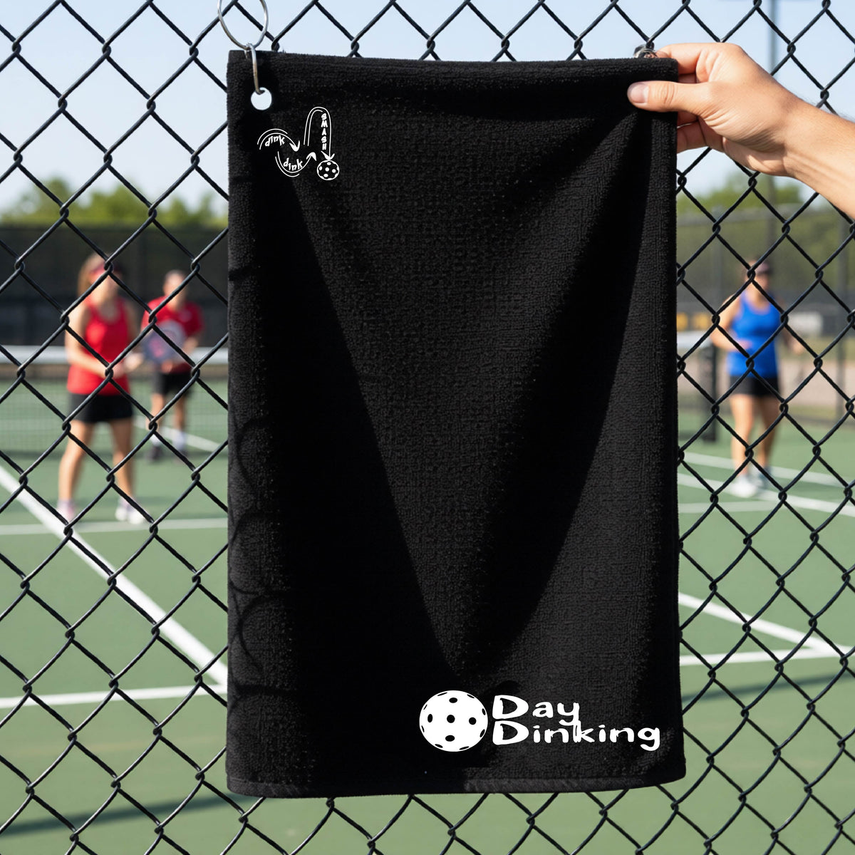 A black terry cloth towel held up by a hand in front of a chain-link fence and pickleball court, with the logo 'DINK DINK SMASH' in white on the top left and a colorful pickleball and the words "Day Dinking" printed near the bottom.