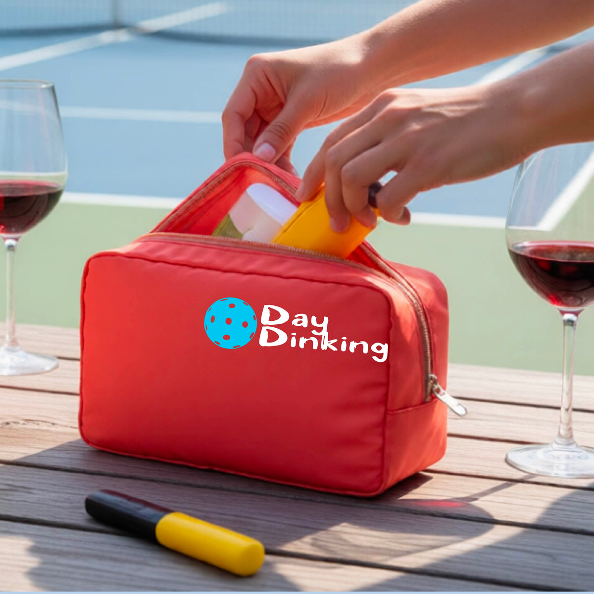 A coral colored zippered cosmetic pouch with a white 'Day Dinking' graphic with a colorful pickleball icon, placed on a wooden table outdoors. 