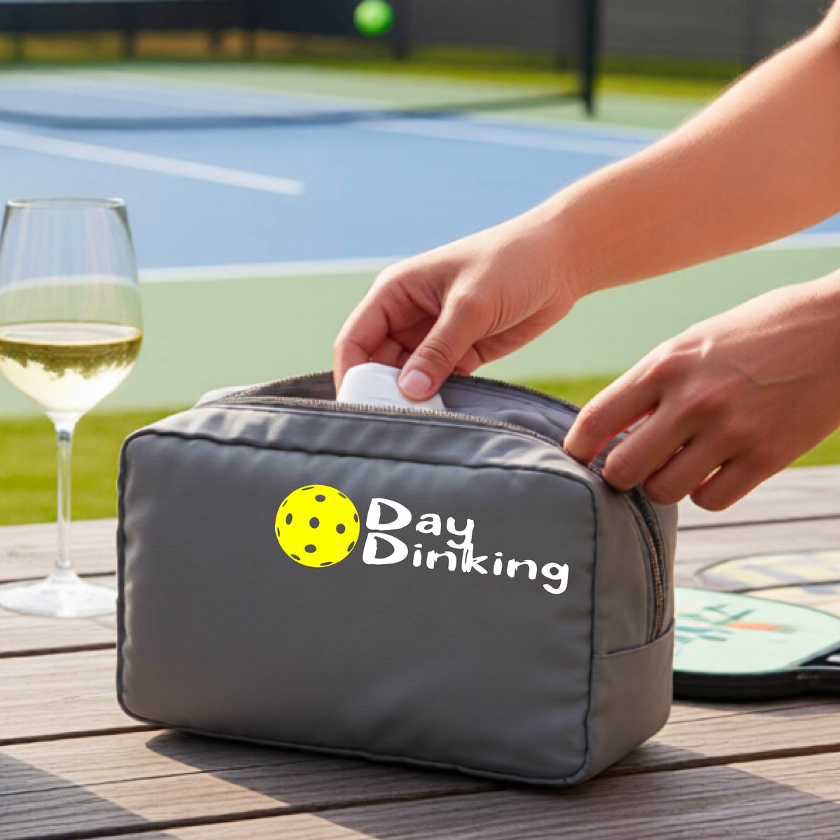 A gray zippered cosmetic pouch with a white 'Day Dinking' graphic with a colorful pickleball icon, placed on a wooden table outdoors. 