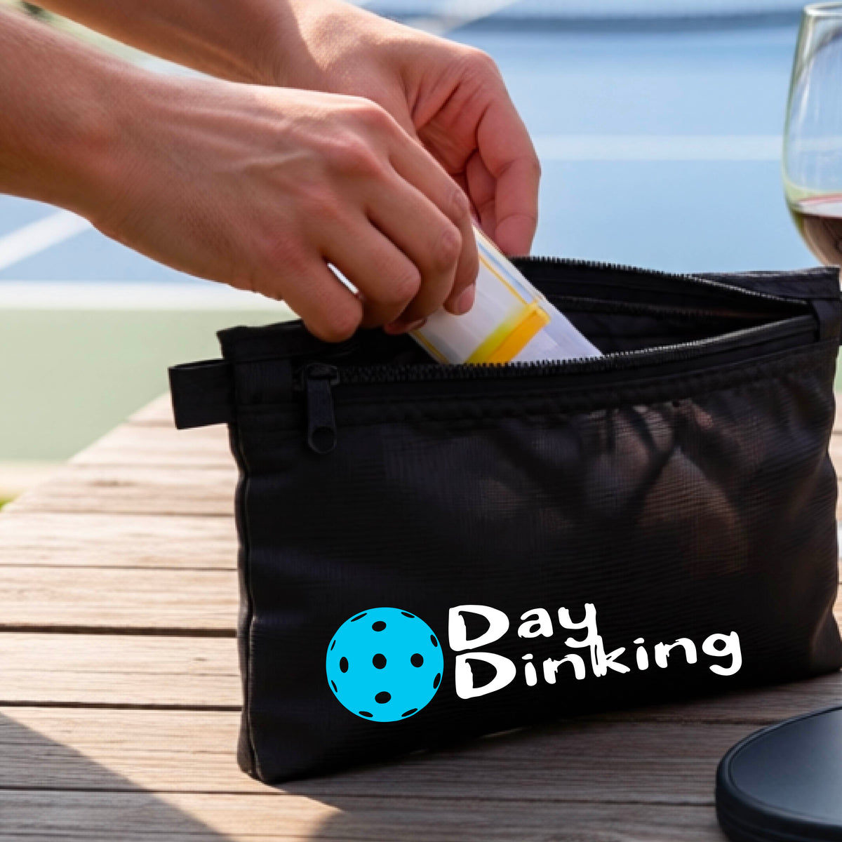 A black zippered cosmetic pouch with a white 'Day Dinking' graphic with a colorful pickleball icon, placed on a wooden table outdoors. 