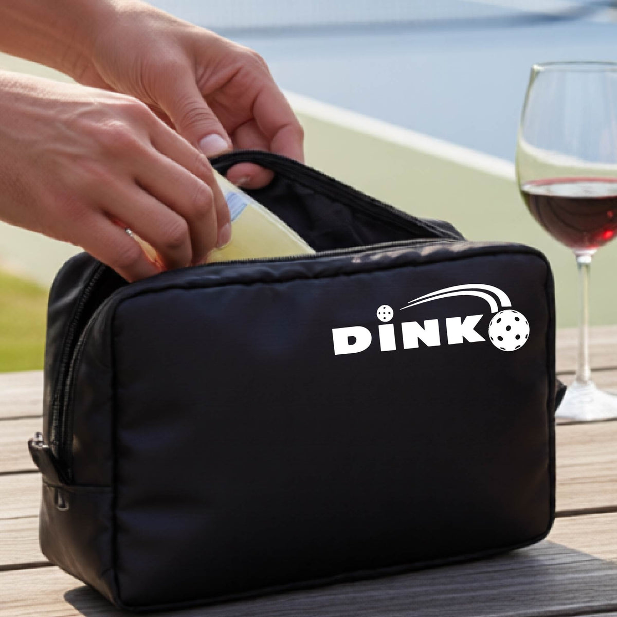 A black zippered cosmetic toiletry bag is shown on a table with a white "DINK" graphic with a white pickleball in flight in the corner with a pickleball court in the background. 