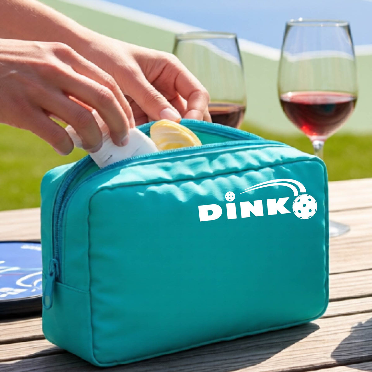 A turquoise zippered cosmetic toiletry bag is shown on a table with a white "DINK" graphic with a white pickleball in flight in the corner with a pickleball court in the background. 
