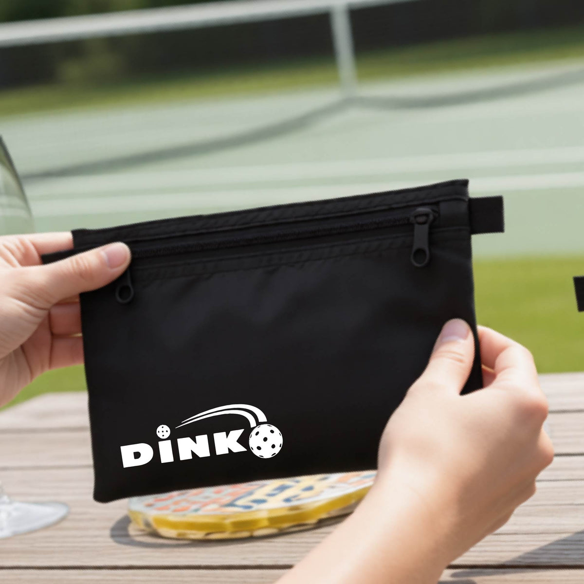 A close up of a black zippered cosmetic bag with a pickleball design of text 'DINK' with a white pickleall in flight, with a pickleball court in the background. 