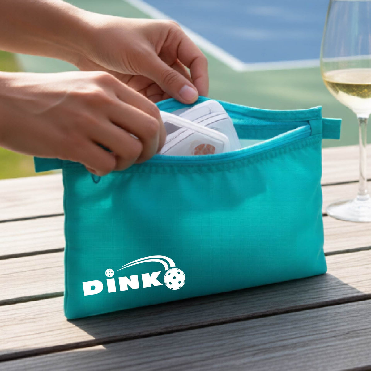 A close up of a aqua blue zippered cosmetic bag with a pickleball design of text 'DINK' with a white pickleall in flight, with a pickleball court in the background. 