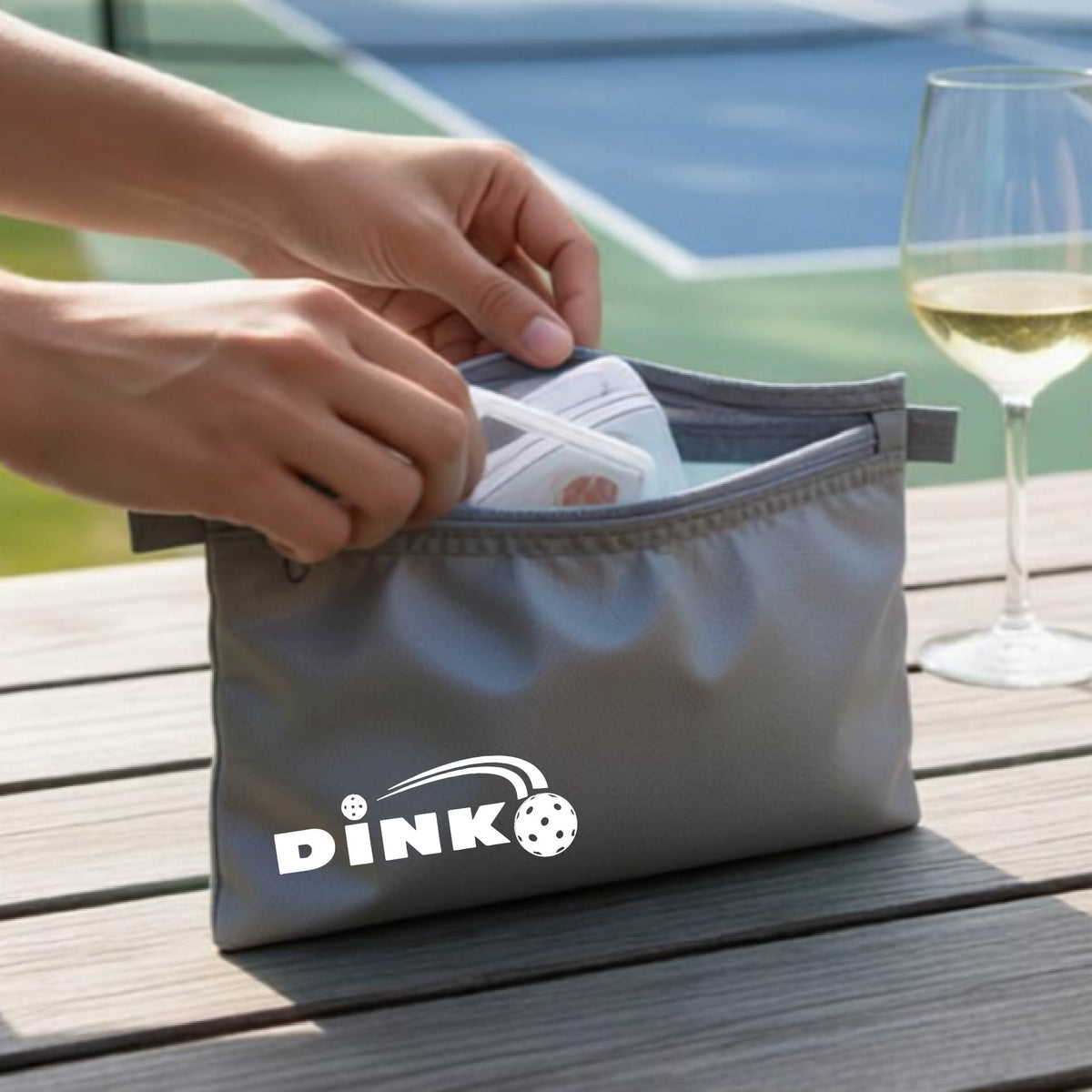 A close up of a gray zippered cosmetic bag with a pickleball design of text 'DINK' with a white pickleall in flight, with a pickleball court in the background. 